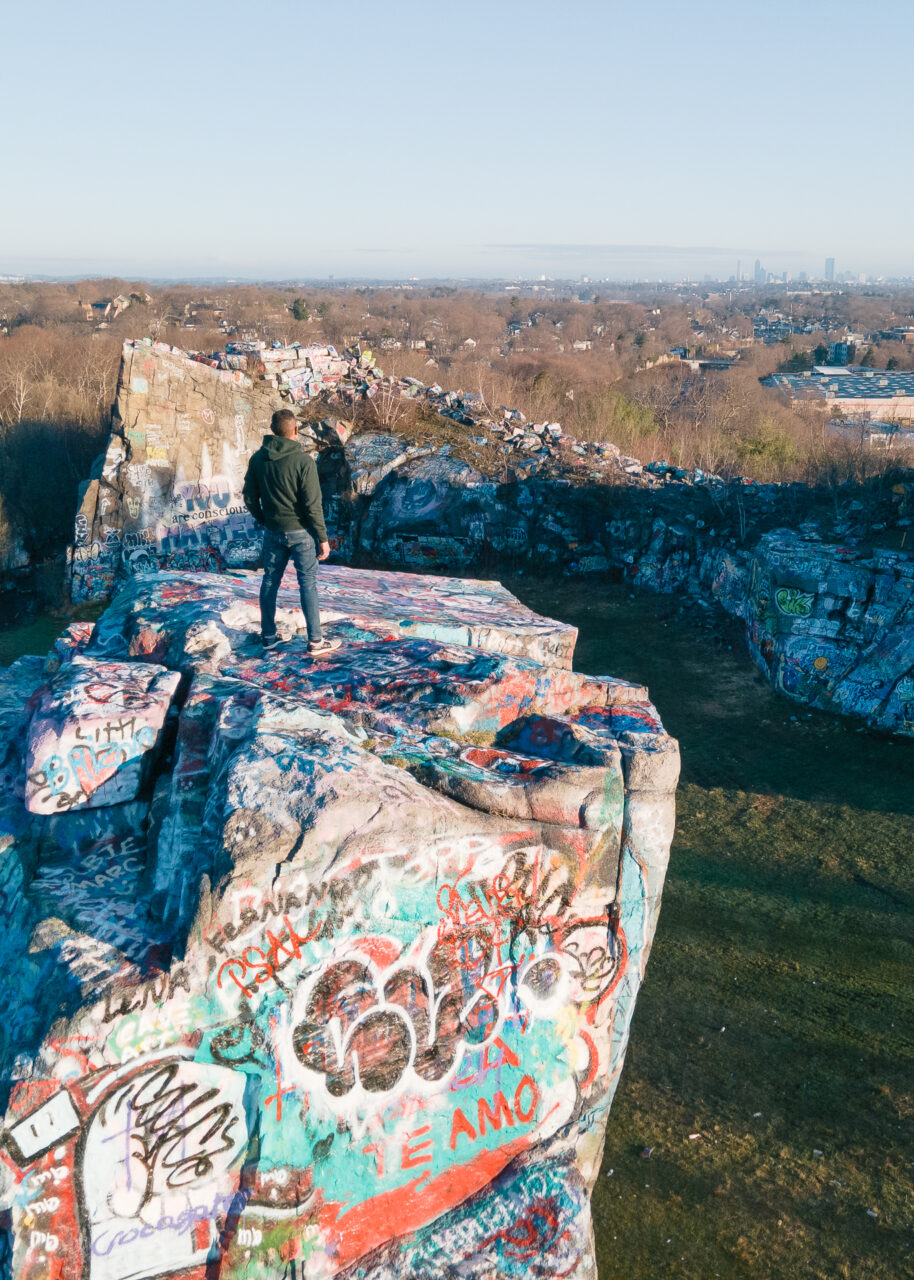 A photo of Lee Costa standing on a rocky, graffiti-covered ledge, gazing toward the Boston skyline.