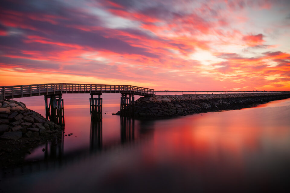 The Plymouth Jetty reflected in the calm waters of Plymouth Harbor during sunrise.