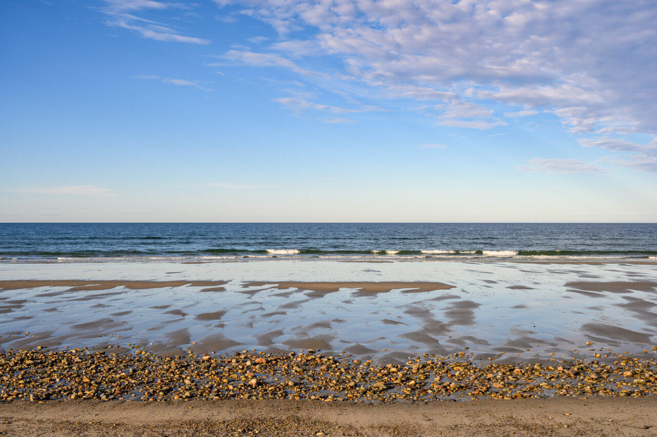 A beach in Duxbury leads to the ocean, where waves crash at the shore under bright blue skies.