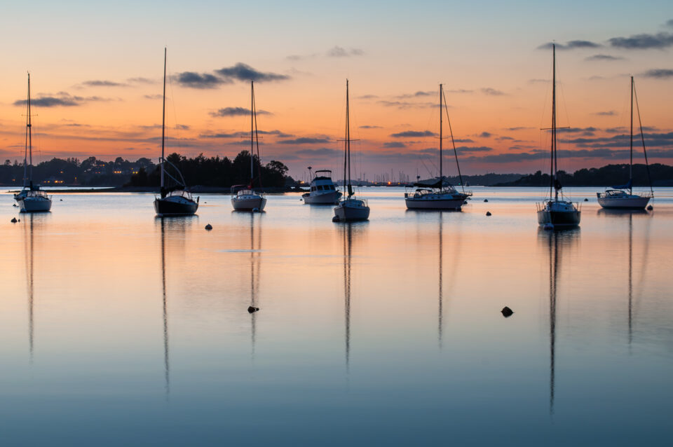 At sunset, boats reflect in the waters of Hingham Harbor, Massachusetts, with vibrant sky colors.