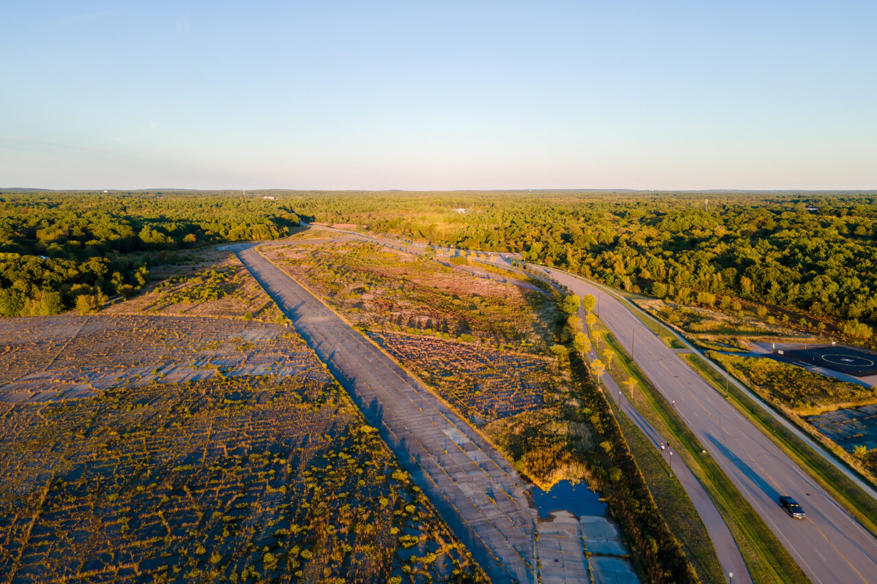 Wide aerial of South Weymouth's cracked runways and weeds, parallel to a new paved road.