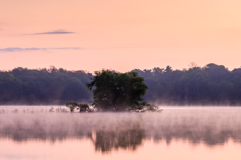 Mist rises over a pond in the morning in Scituate, Massachusetts, with orange skies reflecting in the calm water.