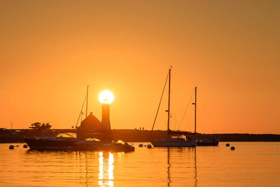 The sun rises above the dome of Scituate Lighthouse in Scituate, Massachusetts, with boats silhouetted against the bright orange sky.