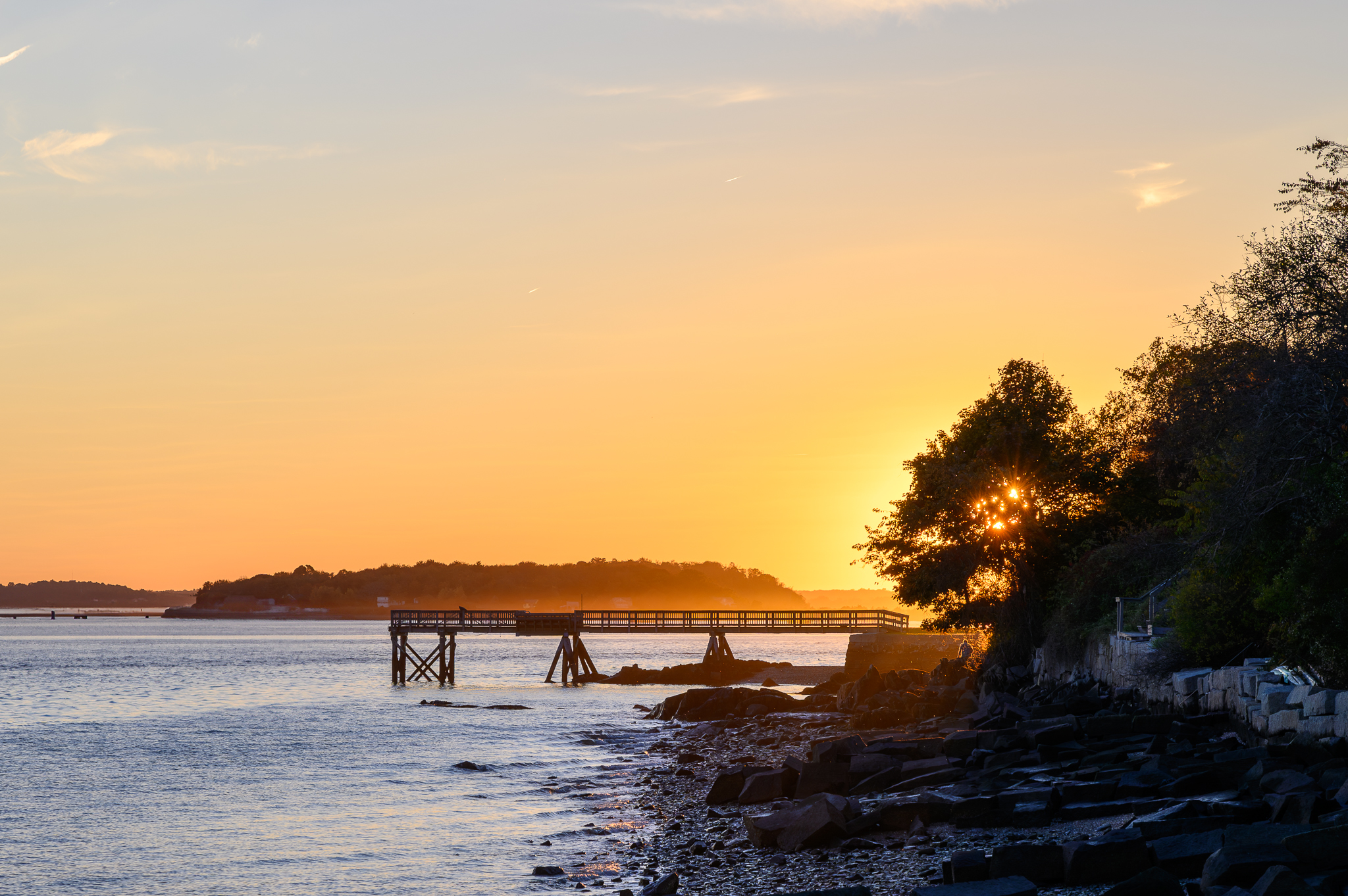 A man sits, hidden among the rocks by a pier, enjoying the sunset.