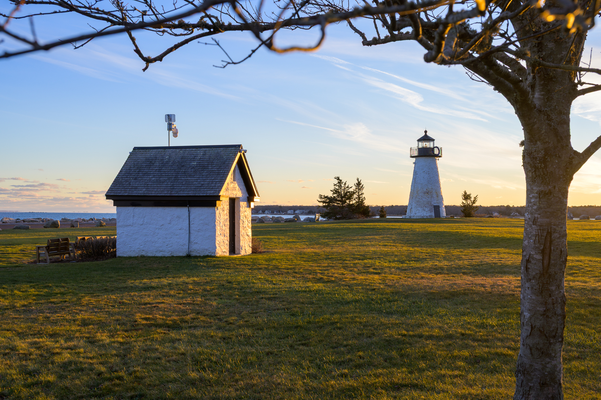 Neds Point Lighthouse, framed by a tree branch with a small house in the background during golden hour.