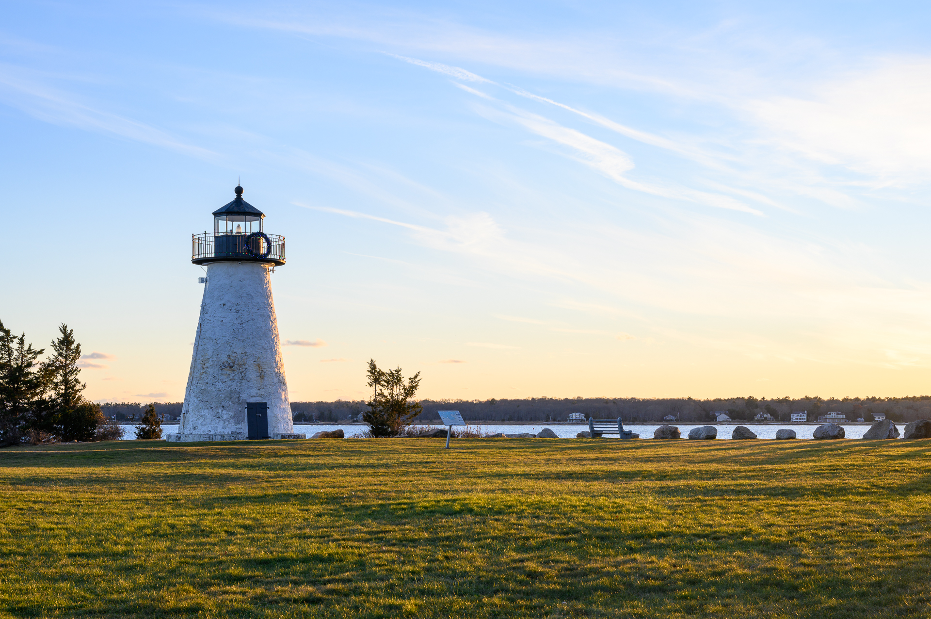 Ned's Point Lighthouse in Mattapoisett bathed in late afternoon light, with warm golden grass stretching toward the harbor and wispy clouds above a sunset sky.