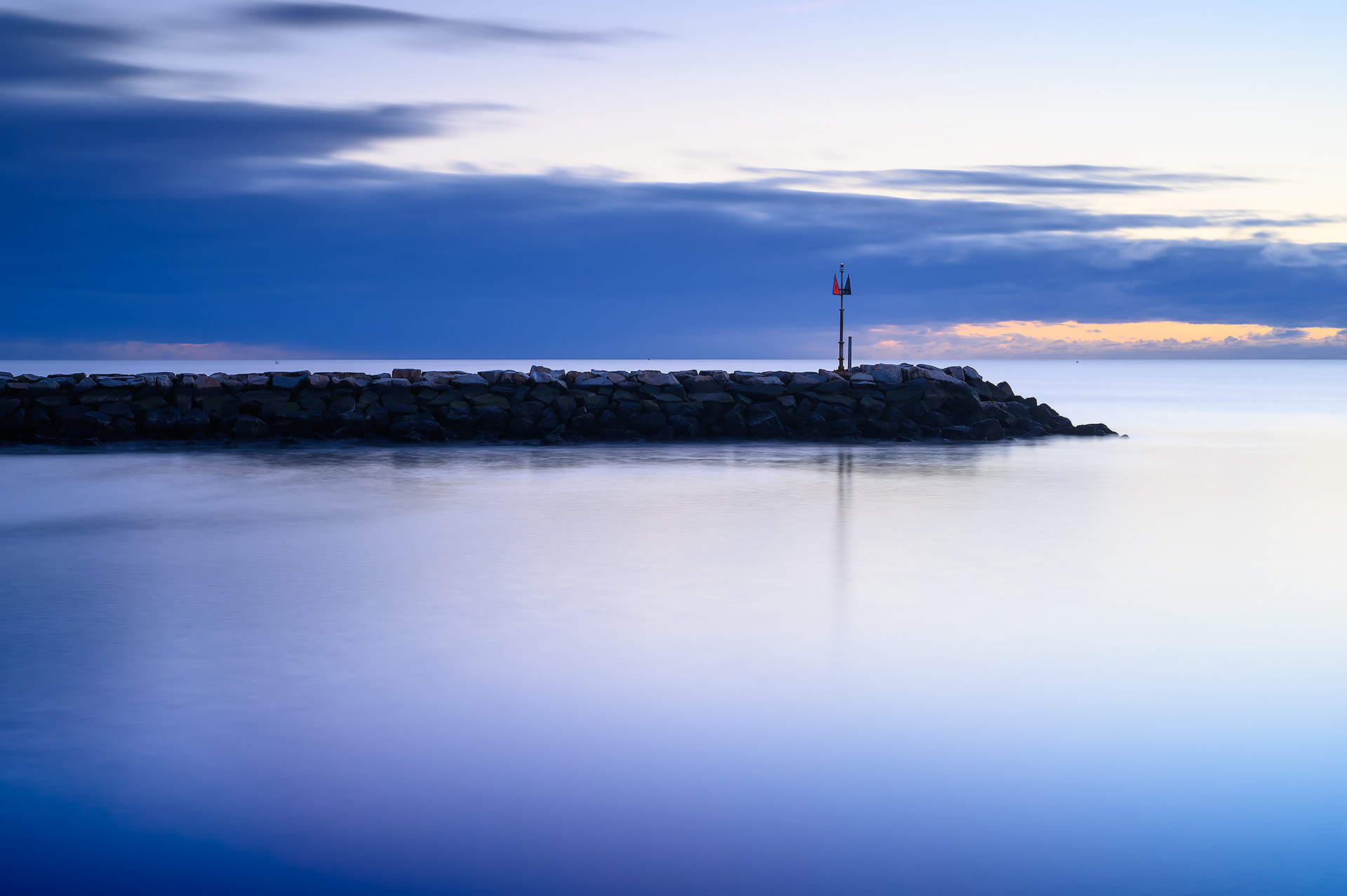 Calm waters surround a jetty at Green Harbor Beach in Marshfield, Massachusetts, as the soft light of sunrise begins to brighten the horizon.