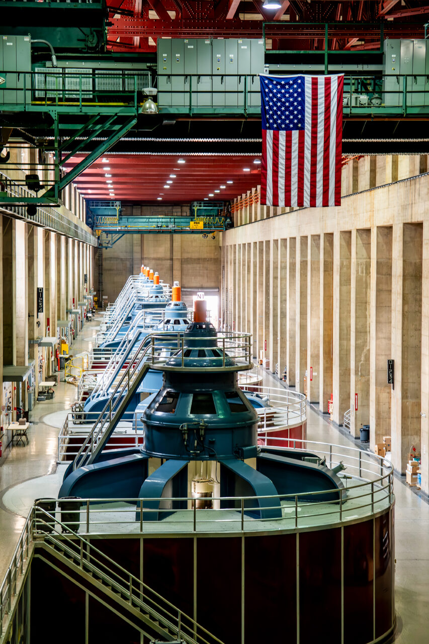 View of the Hoover Dam powerhouse interior with multiple turbines lined up, overhead cranes, and a US flag hanging in the center.