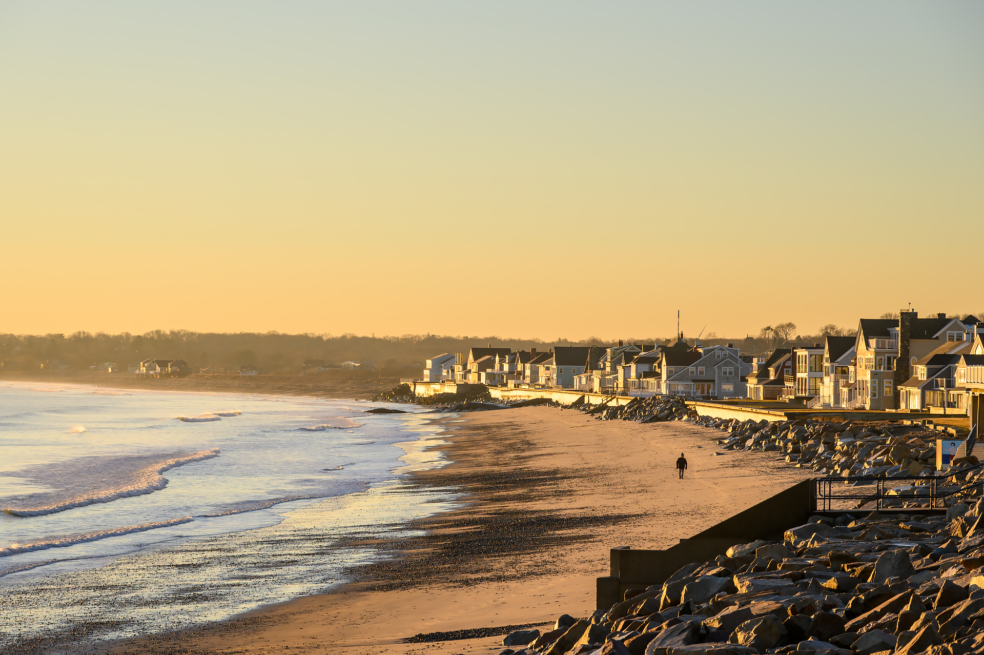 A coastal neighborhood bathed in sunrise light, with a lone person strolling along the sandy beach near the water’s edge.