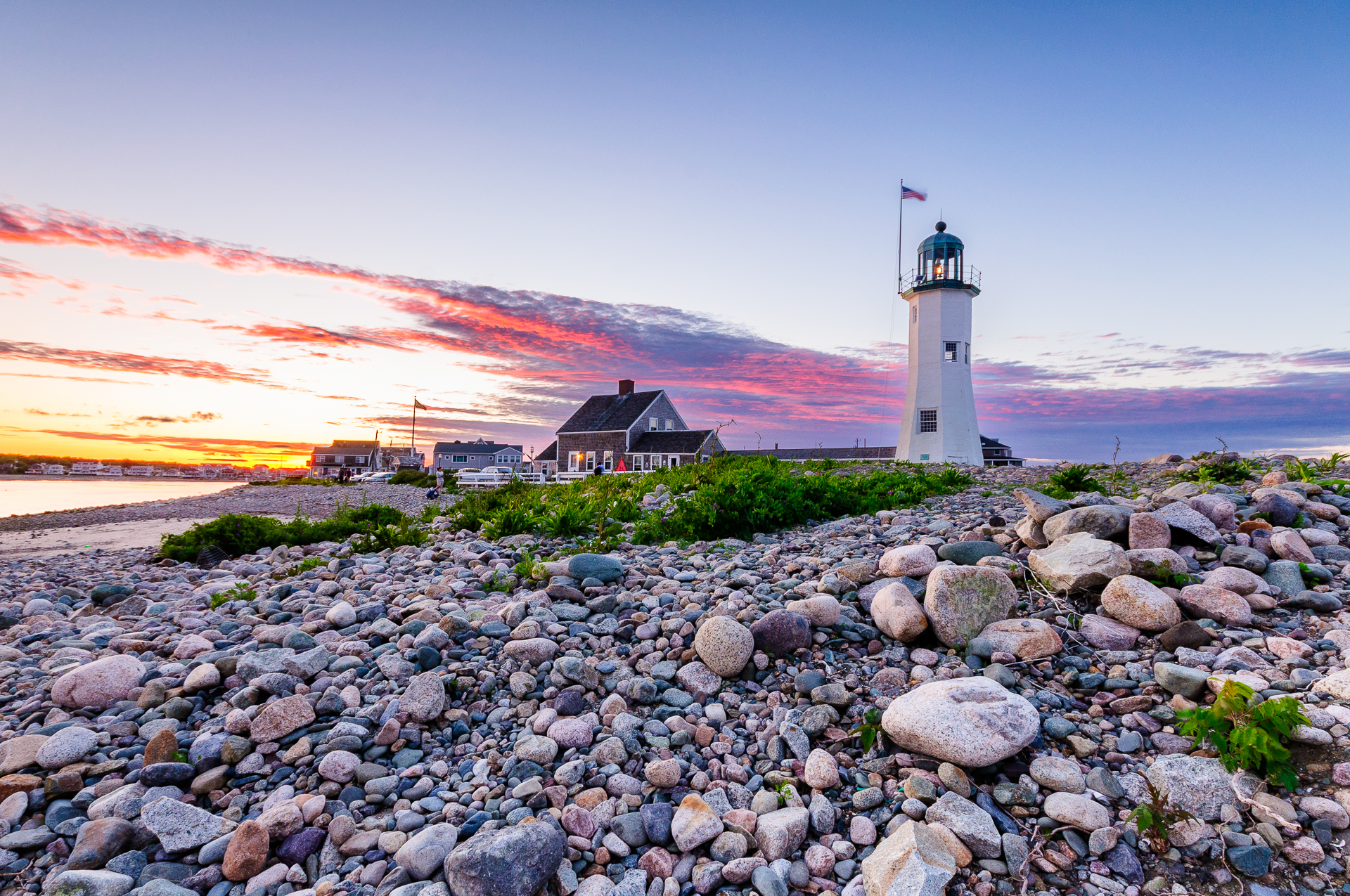 Scituate Lighthouse stands tall at sunset, its light glowing softly as vibrant hues of orange, pink, and purple stretch across the sky above the rocky shoreline.