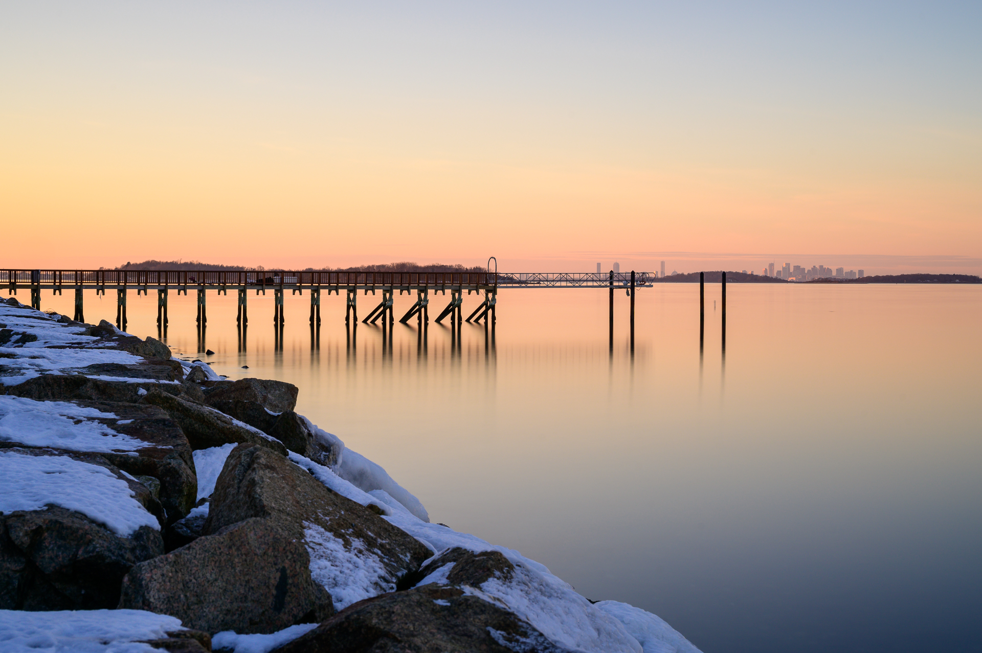 A vibrant sunset over Hingham Harbor with a long pier extending into the water. Snow-covered rocks line the shore in the foreground. The Boston skyline is visible on the horizon.