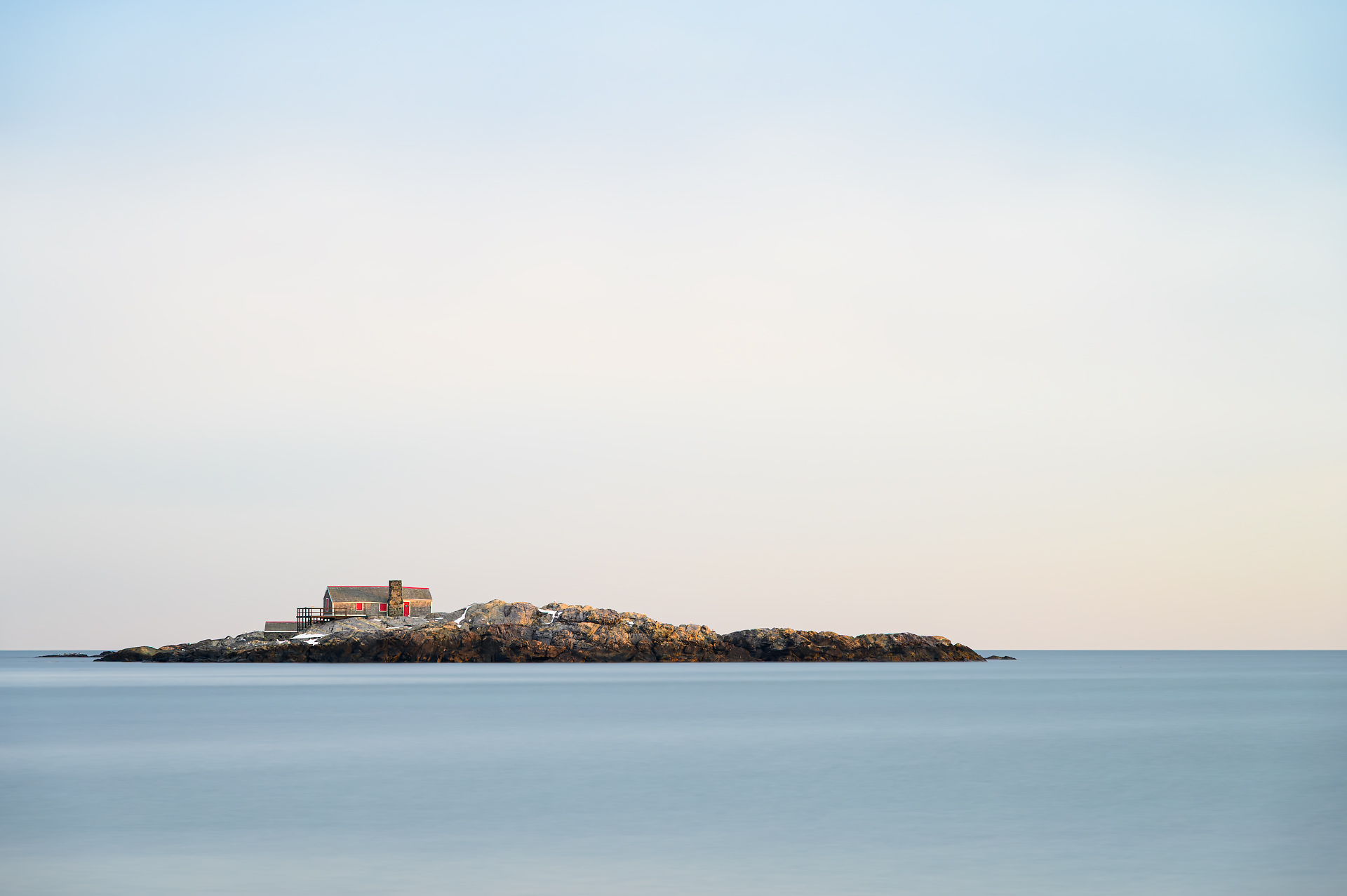 A long-exposure photograph of the Doctor's House on Black Rock Island in Cohasset, Massachusetts. The small, rocky island features a historic cottage with red shutters, standing against a smooth, glass-like sea beneath a soft pastel sky.