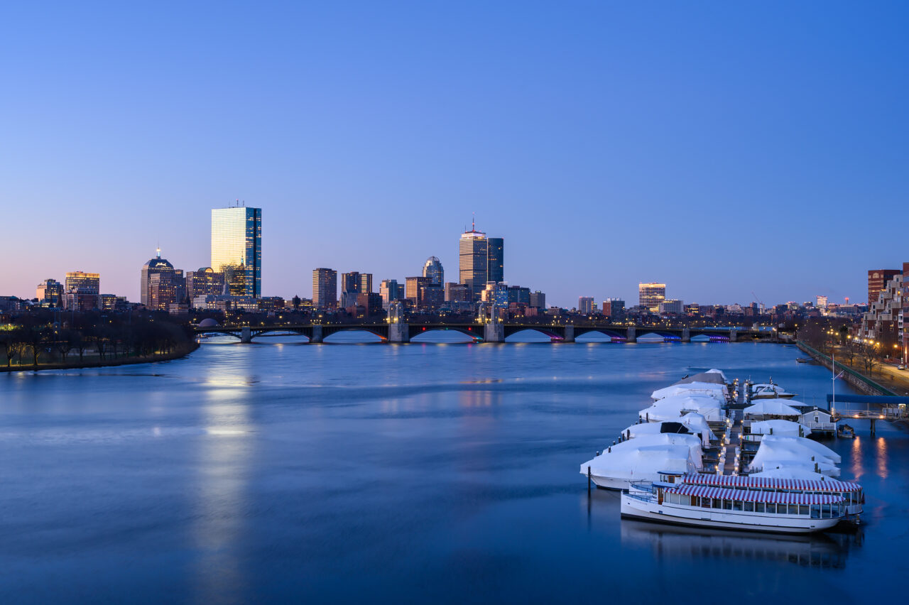 A view of the Boston skyline from a secret spot during the blue hour, with boats in the foreground leading toward the Longfellow Bridge and famous landmarks in the background.