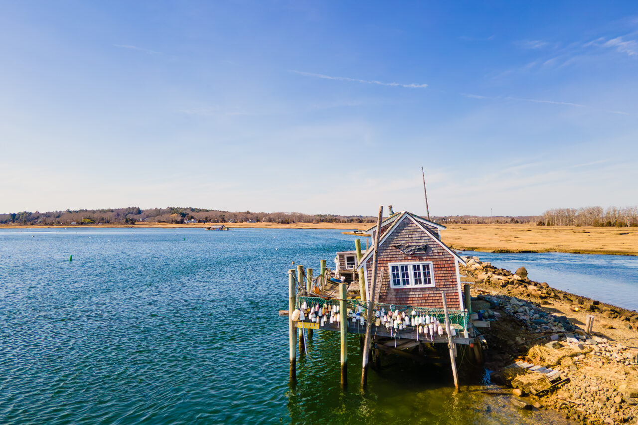 A view of Dauphtucket House by air, a weathered shingle structure on stilts, surrounded by water and marshland.