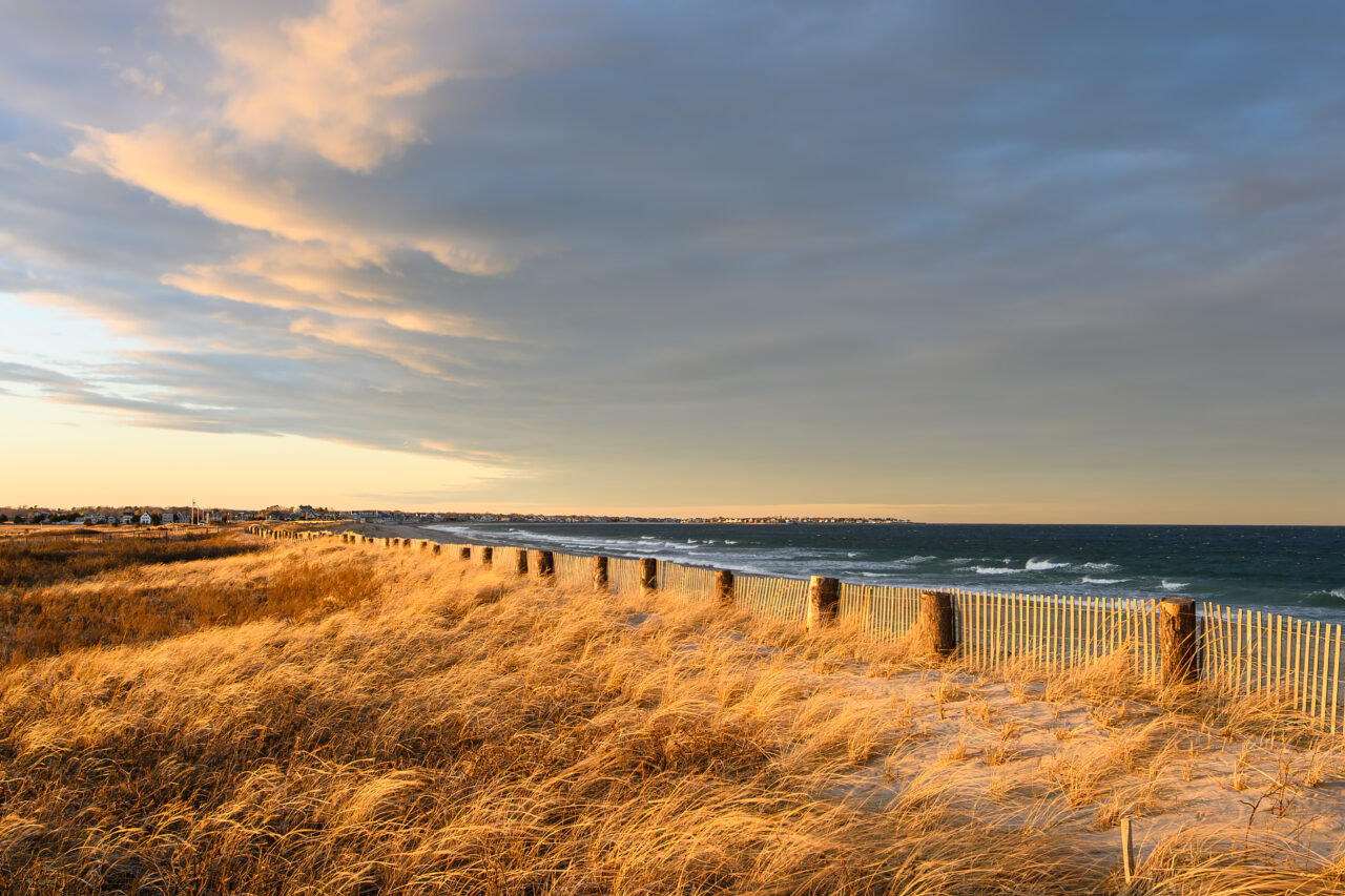 Windswept dunes and golden grasses at Duxbury Beach under a dramatic sunset sky, capturing the raw beauty of the New England coastline.