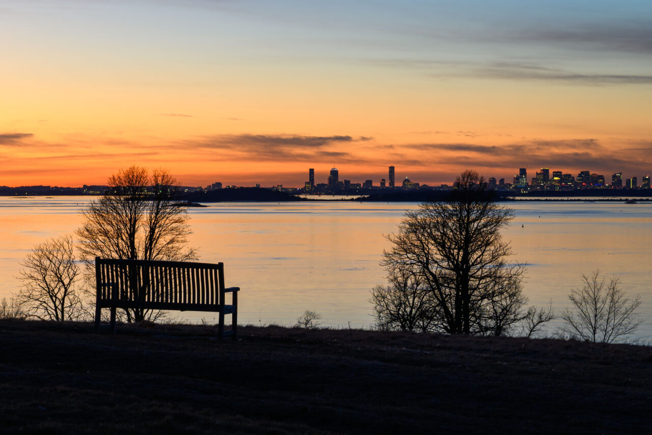 A welcoming park bench looking out toward the glowing Boston skyline during a sunset stroll through World’s End in Hingham.