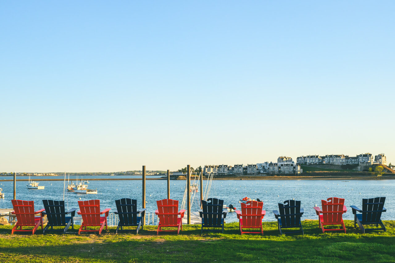 Colorful plastic chairs facing the harbor on a patch of grass, under soft afternoon light in Hull, Massachusetts.