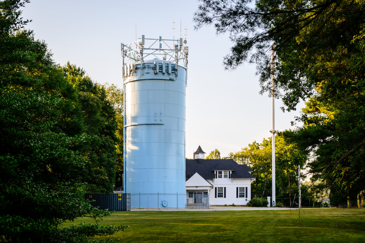 A blue water tower with antennas stands beside a small white house resembling a lighthouse among trees.