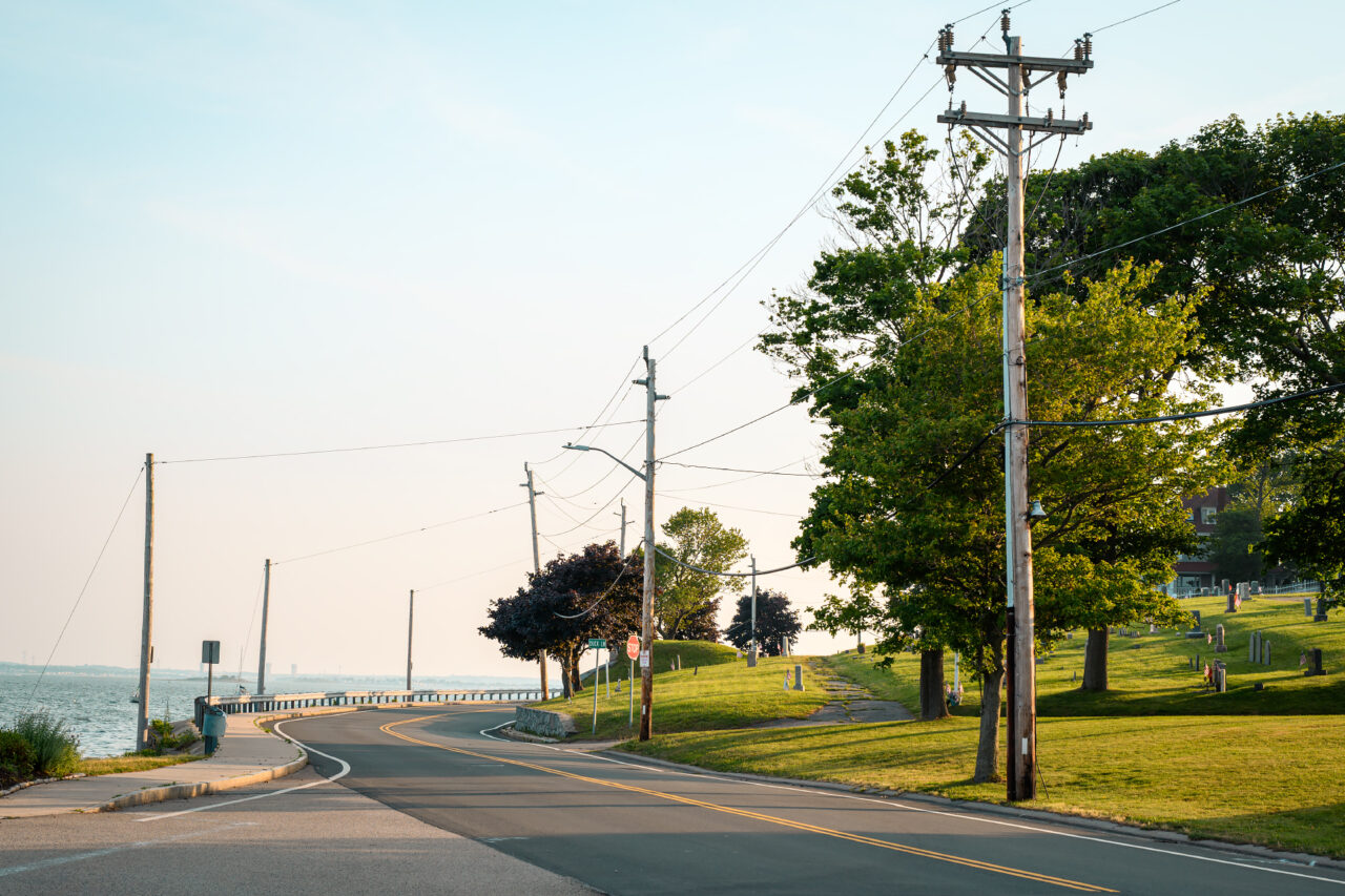 A quiet coastal road curves past a grassy cemetery with mature trees and telephone poles, power lines stretching toward the horizon at sunset.