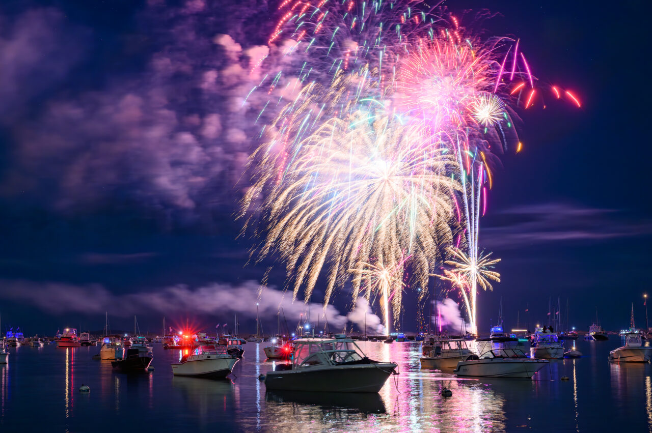 Fireworks display reflecting on the water of Plymouth Harbor, with several boats in the background on the Fourth of July.