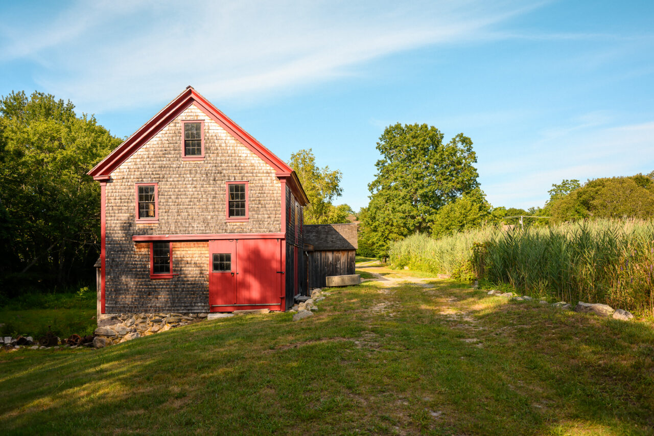 Side view of Hatch Mill building in Marshfield, MA, with a dirt path leading away and tall reeds in the background.