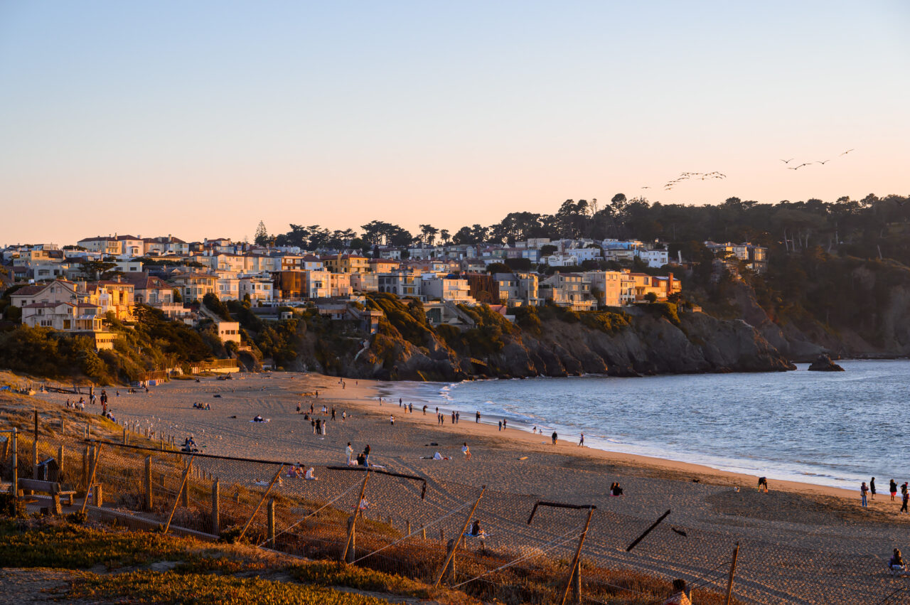 A wide landscape view of Baker Beach in San Francisco at sunset, showing people on the sand and residential buildings on the cliffs overlooking the water.