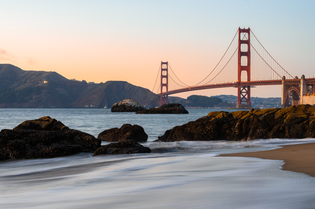 A long-exposure photograph of the Golden Gate Bridge from Baker Beach at sunset, with soft, white ocean waves washing onto the sand in the foreground.