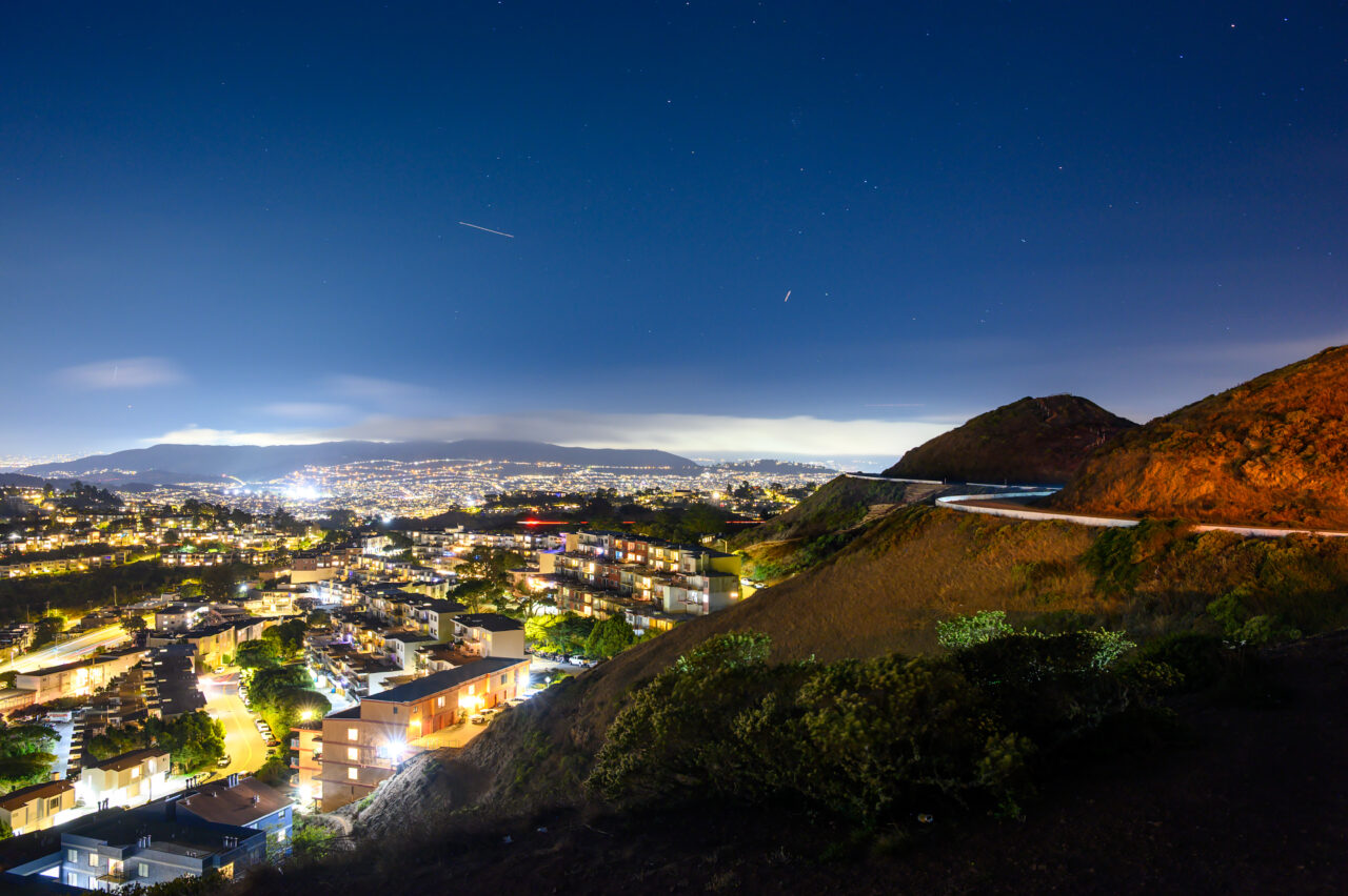 A wide-angle view of the Twin Peaks winding road and the illuminated Bay Area cityscape in the distance.