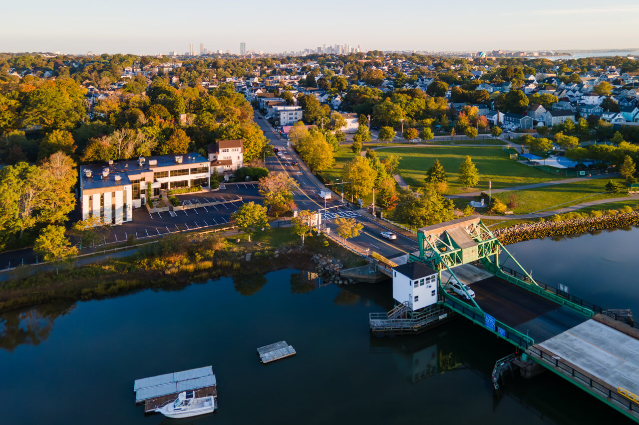 Aerial view of Granite Avenue Drawbridge and neighborhood at sunrise, with road leading to Boston skyline amidst autumn trees.
