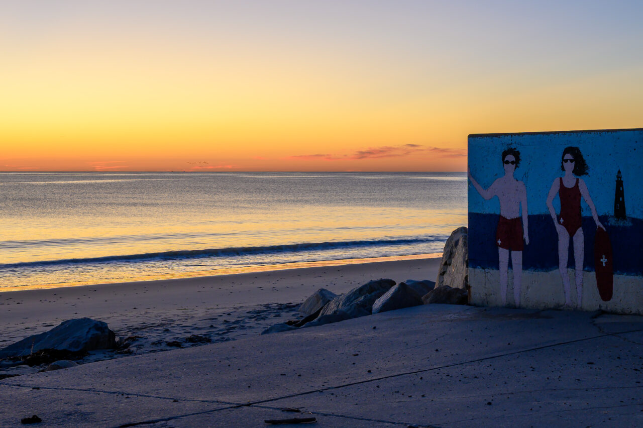 A colorful mural of two lifeguards painted on a concrete wall at Minot Beach during sunrise.