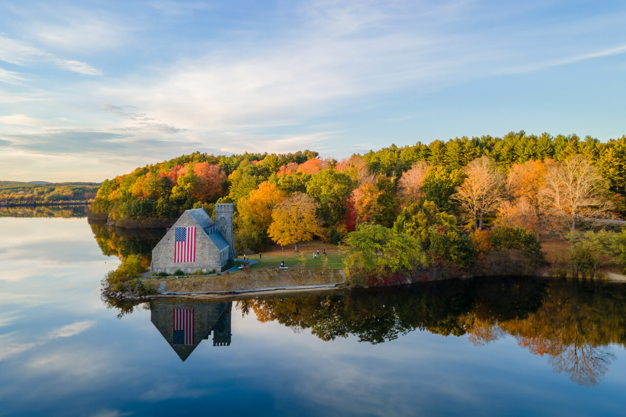 Wide aerial view of the Old Stone Church and its reflection against the colorful autumn forest.