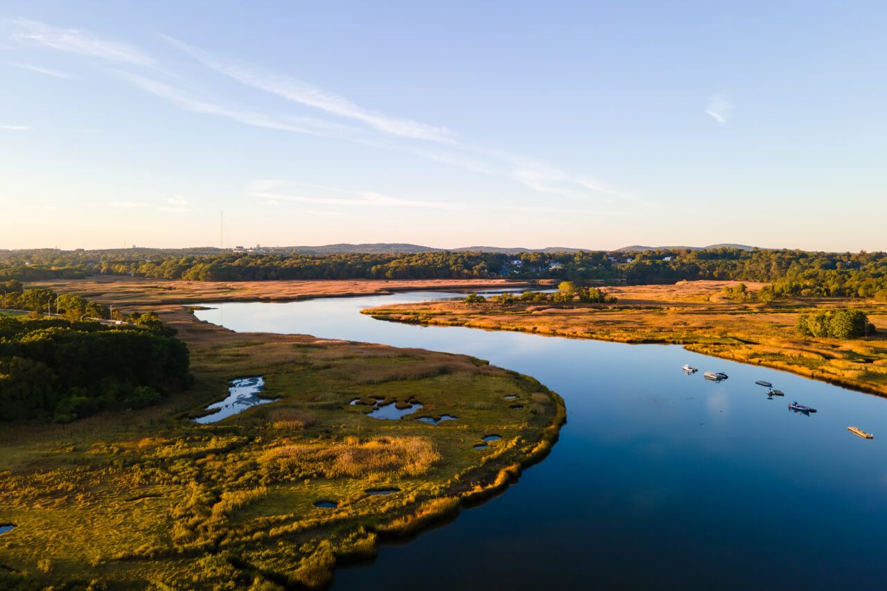Wide aerial view of a winding river and marshland at sunrise, surrounded by sunlit golden grasses and distant autumn trees.