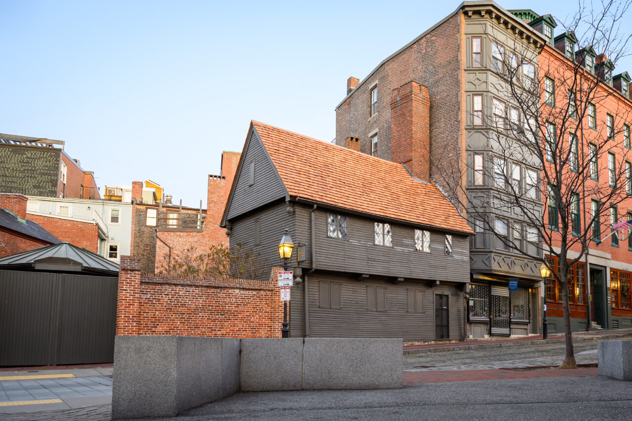 Front view of the Paul Revere House with its dark wood siding and orange roof, flanked by modern red brick buildings.
