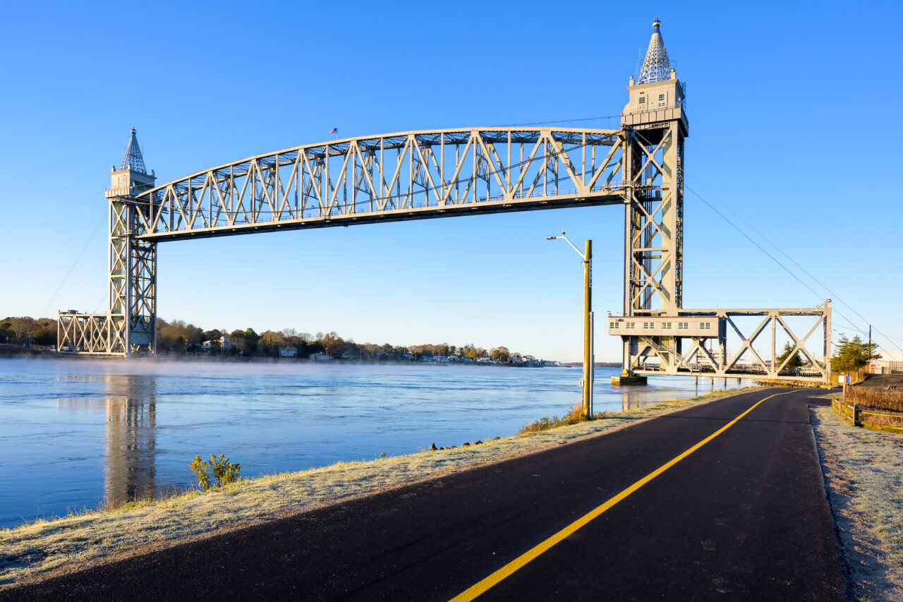 A close view of the Sunrise Cape Cod Canal Railroad Bridge from the bike path, showing the vertical lift structure reflected in the calm water with patches of mist and frost.