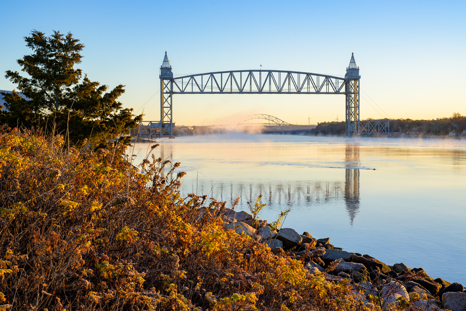 Golden fall foliage lines the rocky edge of the Cape Cod Canal, perfectly framing both the vertical lift Sunrise Cape Cod Canal Railroad Bridge and the arched Bourne Bridge over the morning mist.