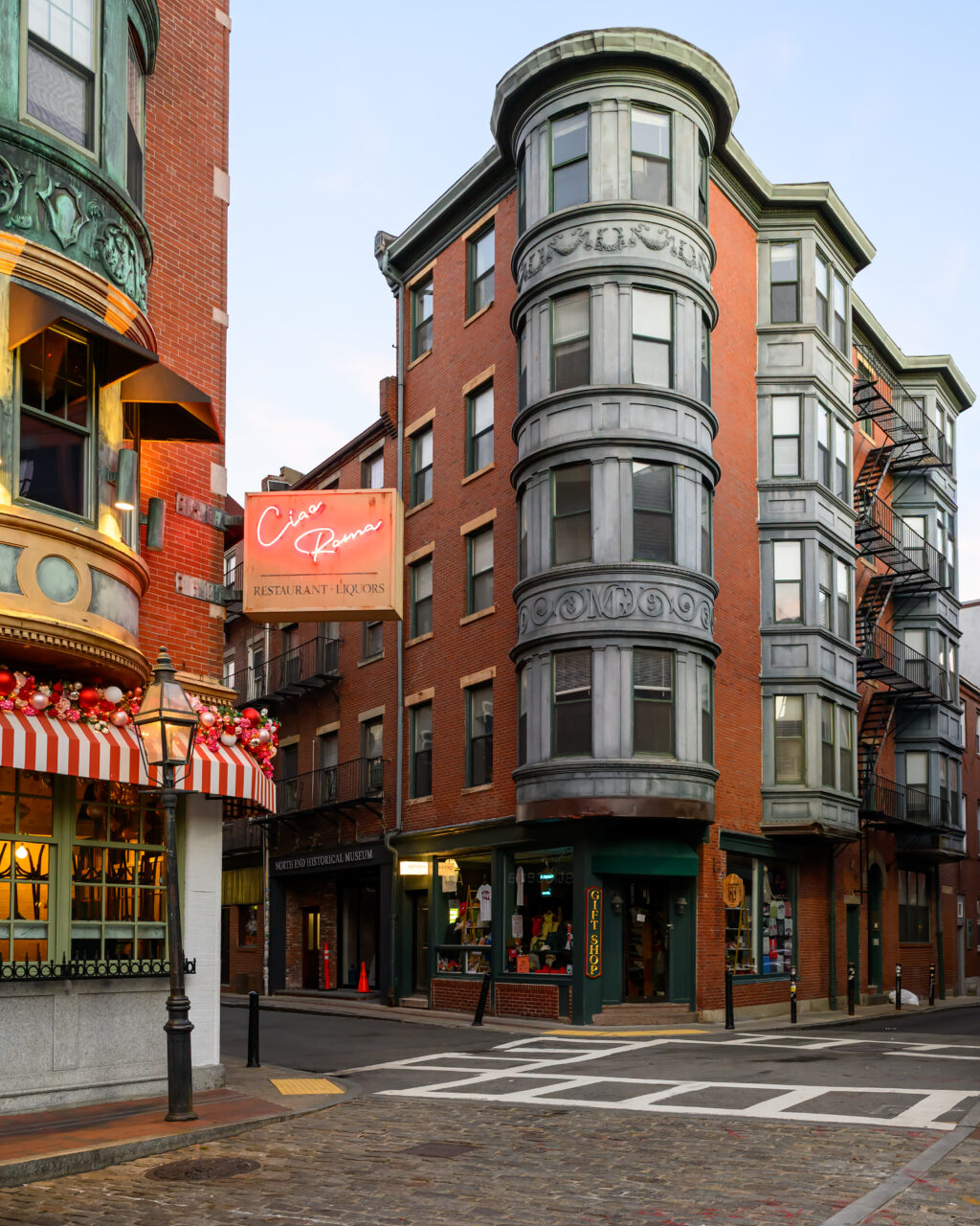 North End street corner with a tall red-brick building, curved metal bay windows, and the illuminated Ciao Roma restaurant sign.