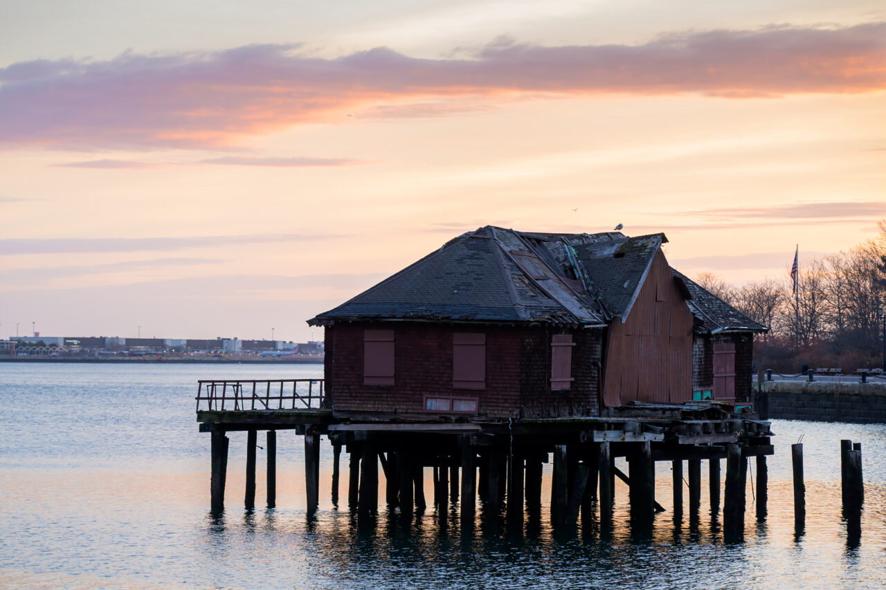 The rustic Northern Avenue bridge tender house at dawn, with the Boston harbor and a distant shoreline visible under a colorful winter solstice sky.