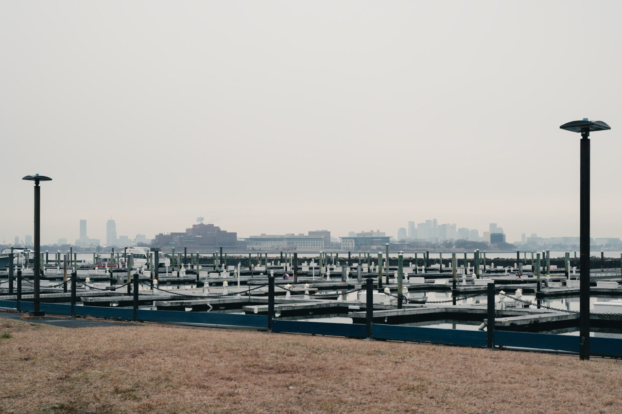 A wide view across dozens of empty boat slips at the marina in Quincy, where the Boston city skyline appears as a soft silhouette on the horizon.