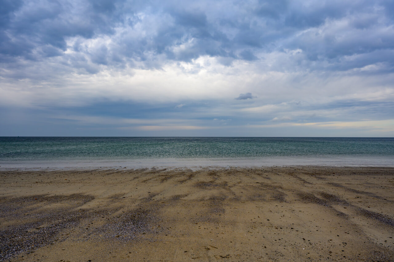 A peaceful view of the tide meeting the sand at Sandwich beach, looking out toward a dark and dramatic winter horizon.