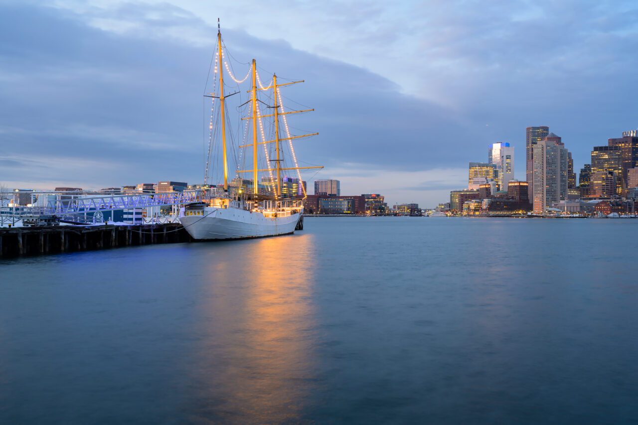 A white sailing ship with golden lights docked at Lewis Mall Wharf marina.