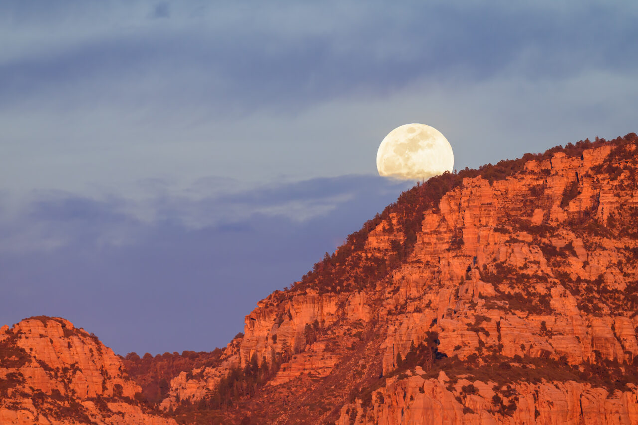 A large, pale full moon rising over the jagged, sun-drenched red rock peaks of Sedona.