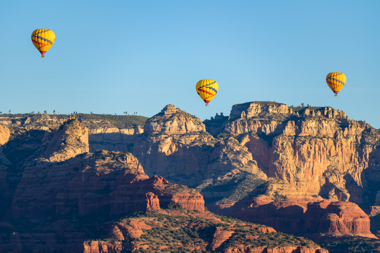Three yellow hot air balloons floating above Sedona's layered red rock canyon walls in early morning light.