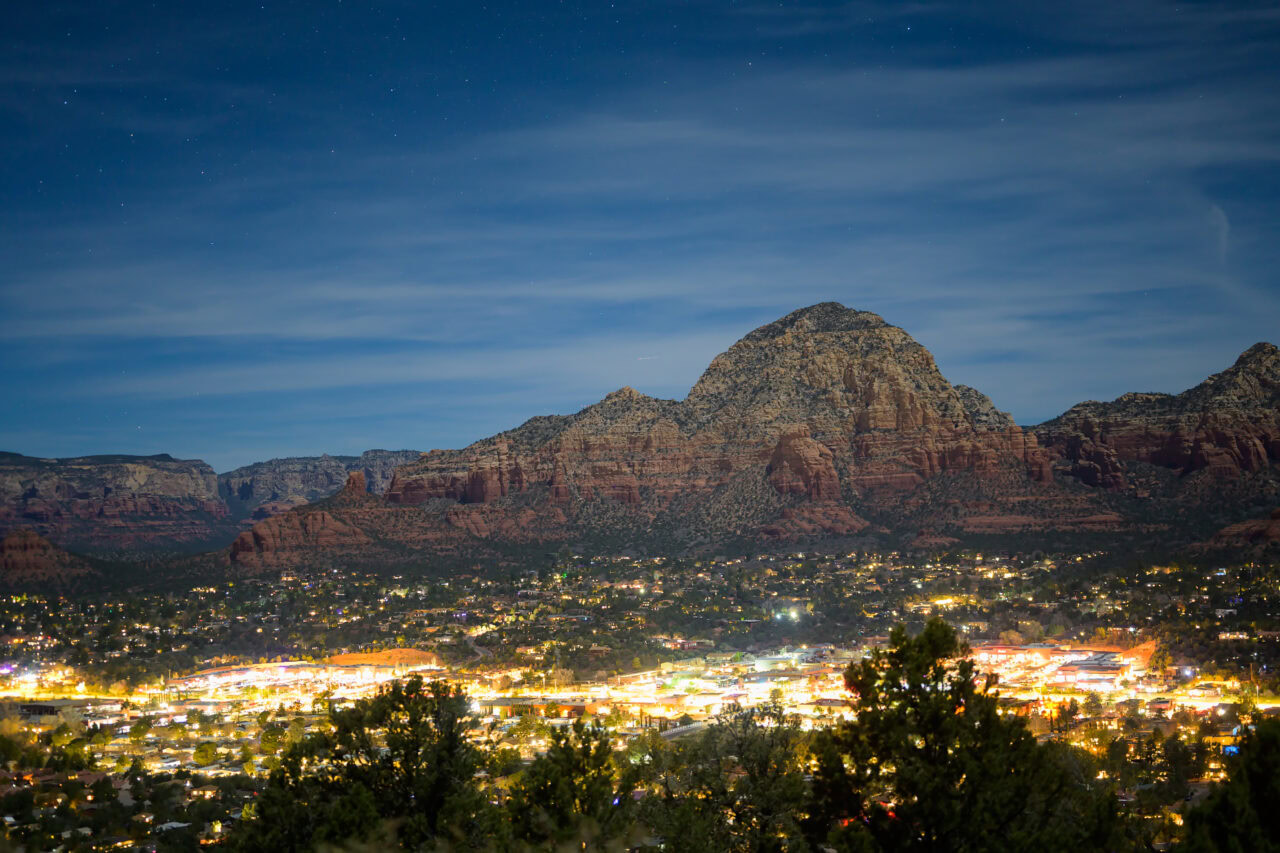 Wide night view from Airport Mesa showing the glowing city of Sedona beneath a starlit sky.