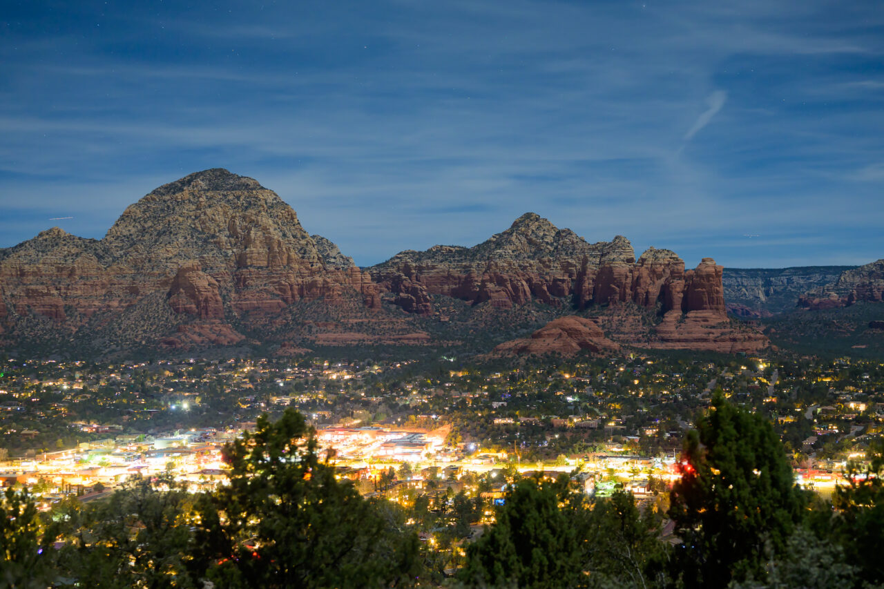 Wide night view from Airport Mesa showing formations including Coffeepot Rock and Sugarloaf Mountain, with the glowing city of Sedona beneath a starlit sky.
