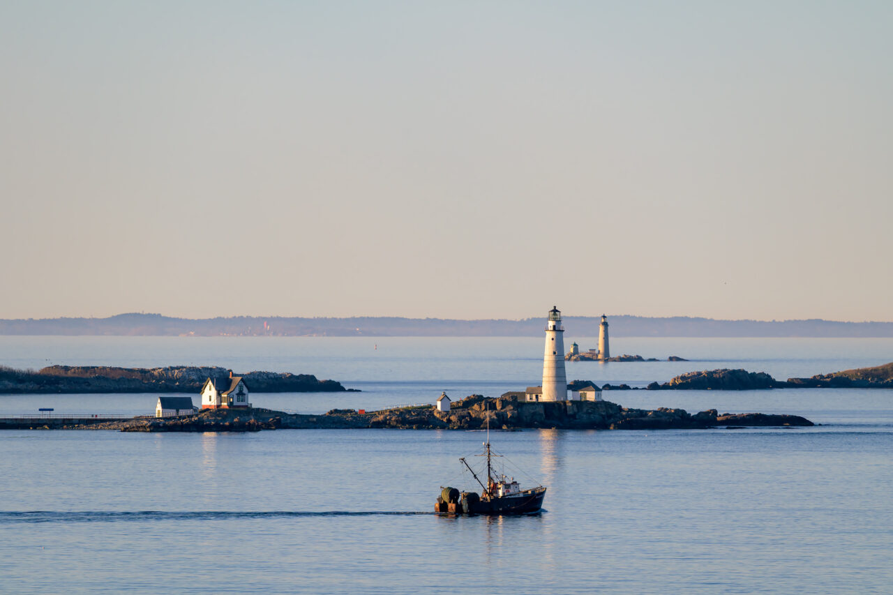 A fishing boat passes before Boston Light on a calm evening in Boston Harbor.