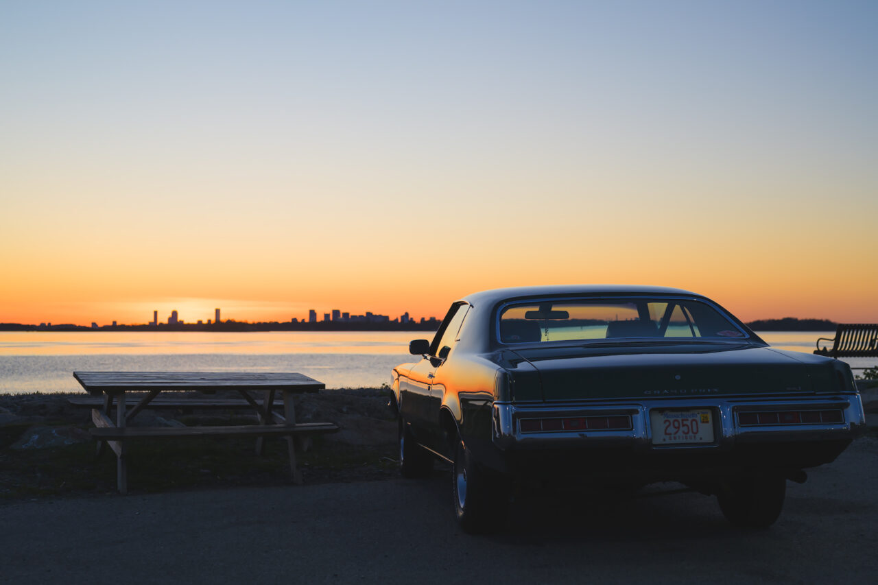 A classic Pontiac Grand Prix parked along the Hull waterfront, with the faint outline of the Boston skyline on the horizon.