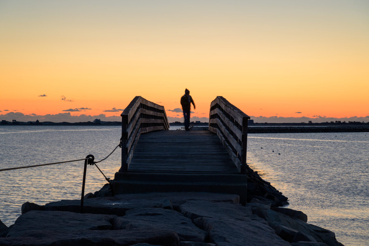 Person walking along Plymouth Jetty at sunrise, silhouetted against calm water and brightening sky.