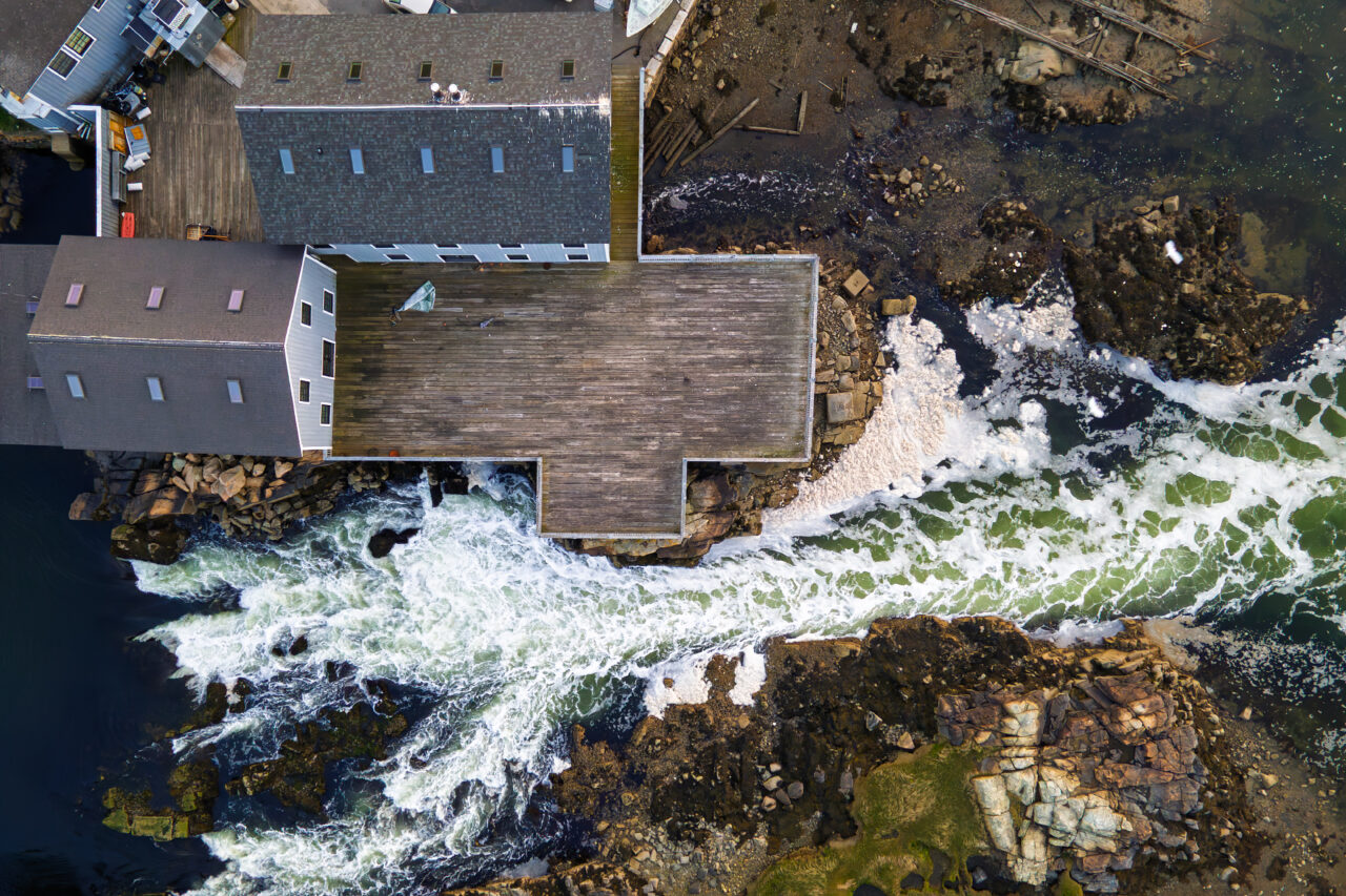 Straight-down aerial view of the Cohasset Lobster Pound featuring a large wooden deck as waves crash over the rocks below in Cohasset Harbor.