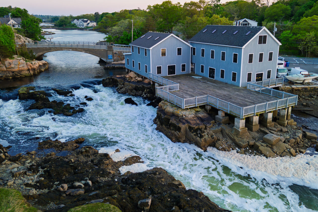 Drone view of the Cohasset Lobster Pound perched over churning tidal rapids at Cohasset Harbor, with a stone arch bridge visible in the background.