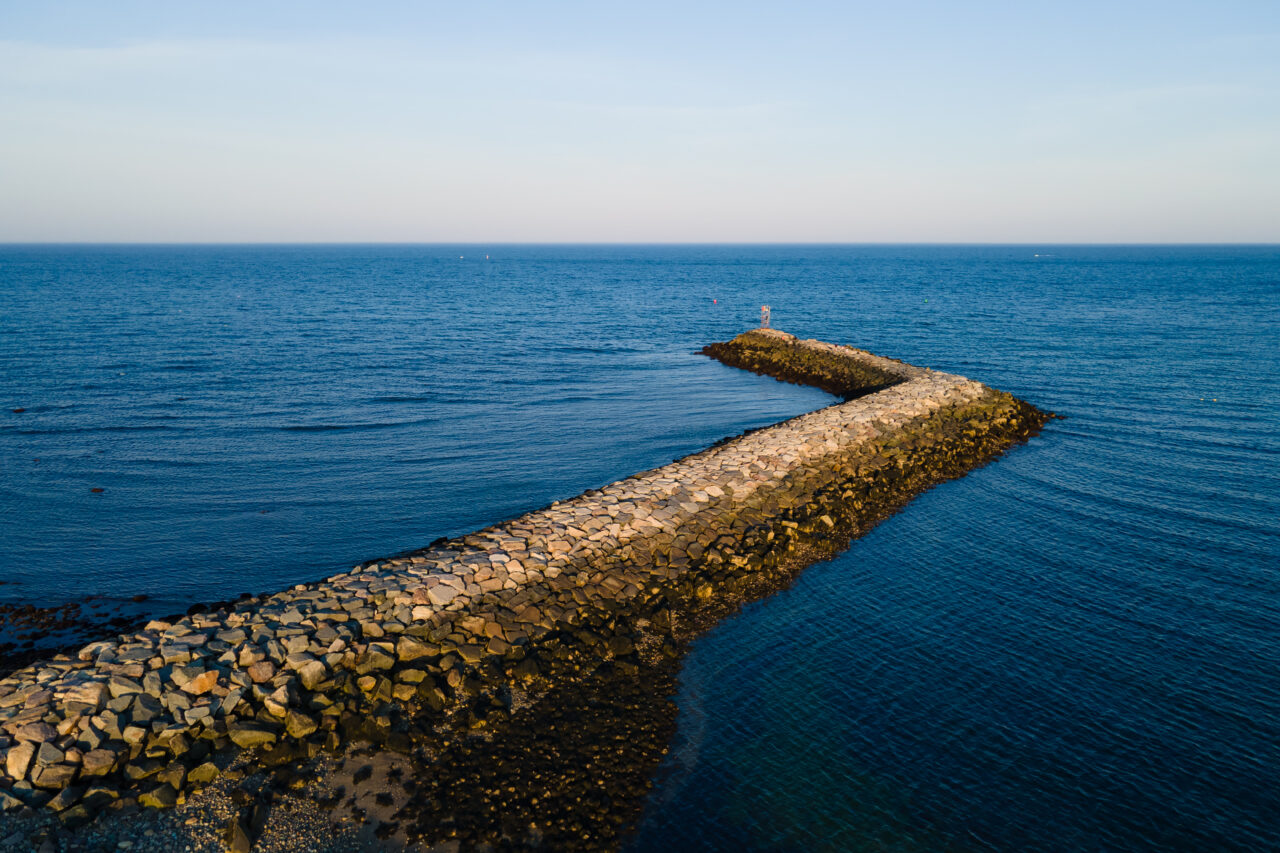 Drone view of the stone breakwater at Scituate Harbor zigzagging out into calm blue water under a clear sky.